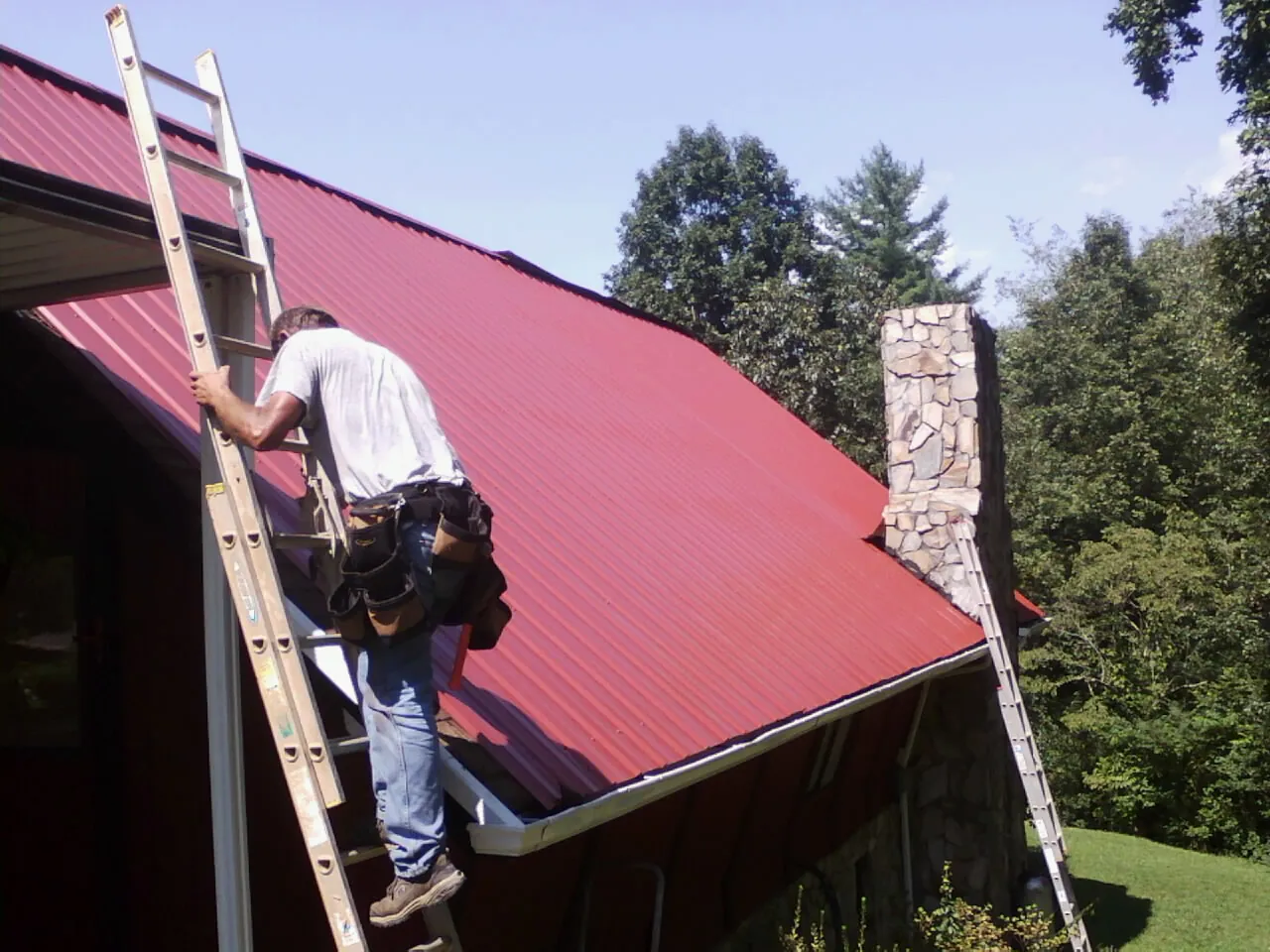Roofer on a ladder beside a red metal roof and stone chimney — Soffit & Fascia Repair by Roofing Brothers in Collinwood